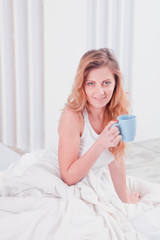 Obraz na płótnie Canvas Good morning and good mood starts with a delicious morning coffee. Portrait of beautiful woman sitting on bed holding a cup of coffee. Vertical image.