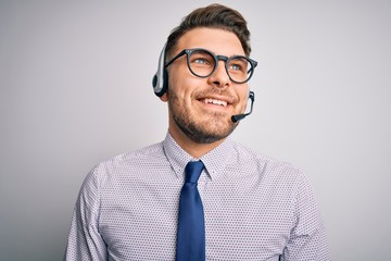 Young call center operator business man with blue eyes wearing glasses and headset looking away to side with smile on face, natural expression. Laughing confident.