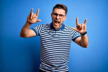Naklejka premium Young man with blue eyes wearing glasses and casual striped t-shirt over blue background shouting with crazy expression doing rock symbol with hands up. Music star. Heavy concept.