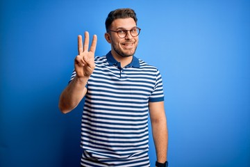 Naklejka premium Young man with blue eyes wearing glasses and casual striped t-shirt over blue background showing and pointing up with fingers number three while smiling confident and happy.