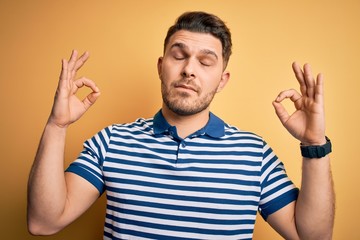 Young man with blue eyes wearing casual striped t-shirt over yellow background relax and smiling with eyes closed doing meditation gesture with fingers. Yoga concept.