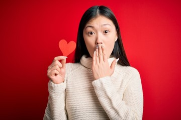 Young asian woman holding romantic red heart paper shape over red isolated background cover mouth with hand shocked with shame for mistake, expression of fear, scared in silence, secret concept