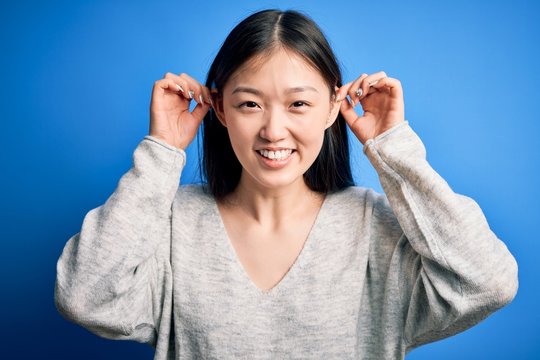 Young beautiful asian woman wearing casual sweater standing over blue isolated background Smiling pulling ears with fingers, funny gesture. Audition problem