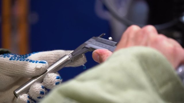 Male welder worker wear protective robe, suit, measures wall thickness of metal gray chiseled part, cut piece of tube with special ruler. Man holds in his right hand measuring tool, caliper close up. 