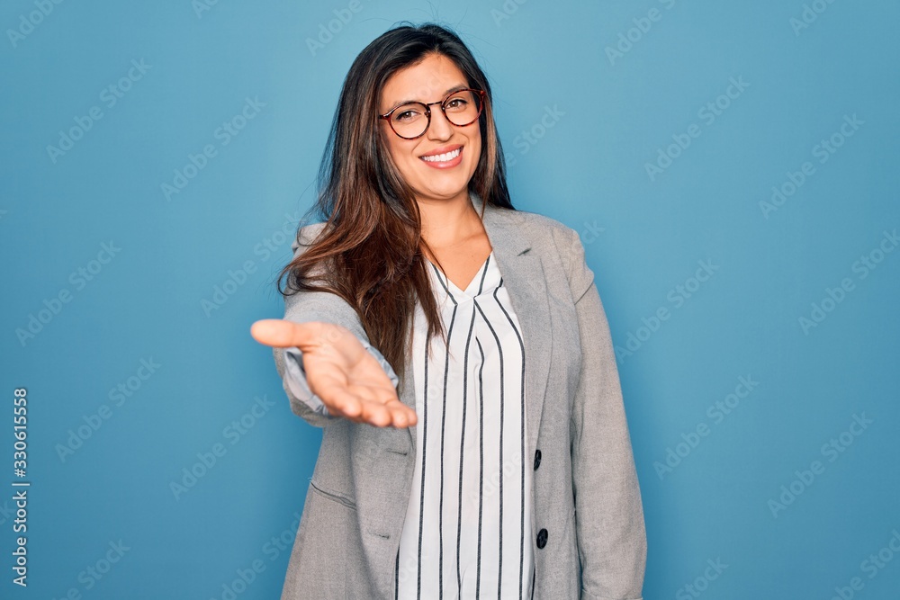 Wall mural young hispanic business woman wearing glasses standing over blue isolated background smiling cheerfu