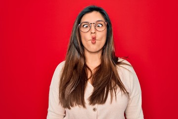 Young hispanic smart woman wearing glasses standing over red isolated background making fish face with lips, crazy and comical gesture. Funny expression.