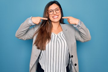 Young hispanic business woman wearing glasses standing over blue isolated background smiling cheerful showing and pointing with fingers teeth and mouth. Dental health concept.