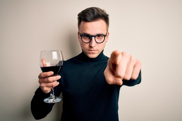 Young handsome caucasian man drinking an alcoholic glass of red wine over isolated background pointing with finger to the camera and to you, hand sign, positive and confident gesture from the front