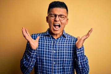 Fototapeta premium Young handsome latin man wearing casual shirt and glasses over yellow background celebrating mad and crazy for success with arms raised and closed eyes screaming excited. Winner concept