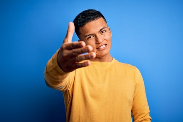Young handsome latin man wearing yellow casual sweater over isolated blue background smiling friendly offering handshake as greeting and welcoming. Successful business.