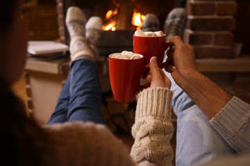 Couple with cups of delicious cocoa resting near fireplace at home, closeup. Winter vacation