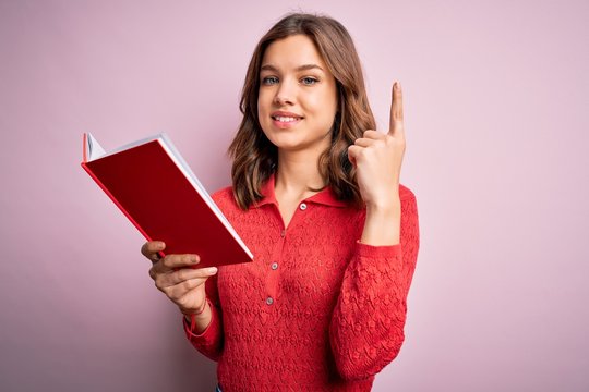 Young blonde student girl reading a book over pink  isolated background surprised with an idea or question pointing finger with happy face, number one