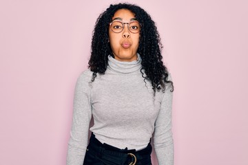 Young african american woman wearing turtleneck sweater and glasses over pink background puffing cheeks with funny face. Mouth inflated with air, crazy expression.