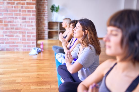 Young beautiful group of sportswomen smiling happy practicing yoga. Sitting doing prayer pose at gym