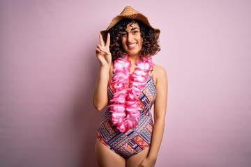 Young beautiful arab woman on vacation wearing swimsuit and hawaiian lei flowers smiling with happy face winking at the camera doing victory sign. Number two.