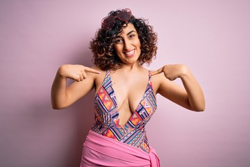 Young beautiful arab woman on vacation wearing swimsuit and sunglasses over pink background looking...