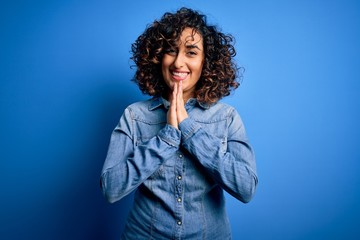 Fototapeta na wymiar Young beautiful curly arab woman wearing casual denim shirt standing over blue background praying with hands together asking for forgiveness smiling confident.