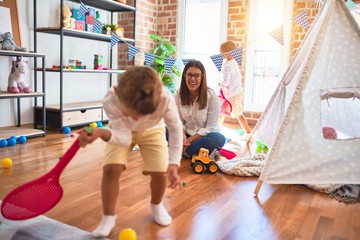 Beautiful teacher and toddlers playing tennis using racket around lots of toys at kindergarten