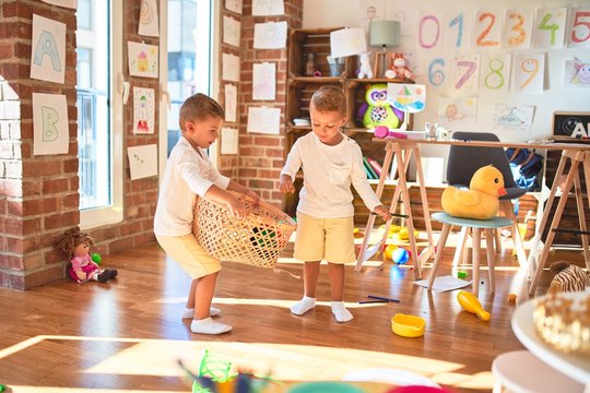 Adorable blonde twins playing basketball using wicker basket and ball around lots of toys at kindergarten