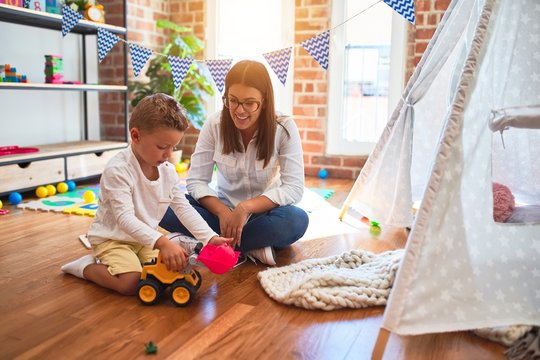 Beautiful teacher and toddler playing with tractor around lots of toys at kindergarten