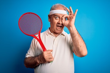 Middle age hoary sportsman playing tennis using racket over isolated blue background with happy face smiling doing ok sign with hand on eye looking through fingers