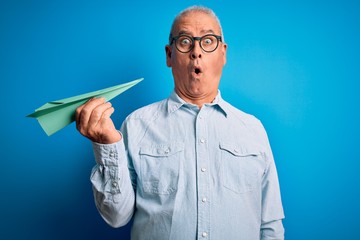 Middle age handsome hoary man holding paper airplane over isolated blue background scared in shock...