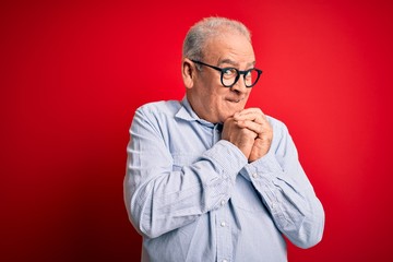 Middle age handsome hoary man wearing casual striped shirt and glasses over red background laughing nervous and excited with hands on chin looking to the side