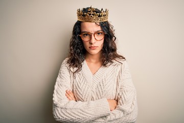 Young beautiful woman with curly hair wearing golden queen crown over white background skeptic and nervous, disapproving expression on face with crossed arms. Negative person.