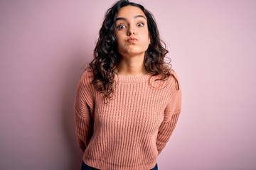 Obraz premium Young beautiful woman with curly hair wearing casual sweater over isolated pink background puffing cheeks with funny face. Mouth inflated with air, crazy expression.