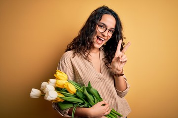 Naklejka premium Young beautiful romantic woman with curly hair holding bouquet of yellow tulips smiling with happy face winking at the camera doing victory sign with fingers. Number two.