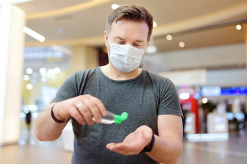 Man wearing disposable medical face mask makes disinfection of hands with sanitizer in airport,...