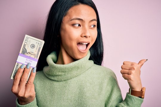 Young beautiful chinese woman holding dollars standing over isolated pink background pointing and showing with thumb up to the side with happy face smiling