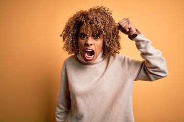 Young beautiful african american woman wearing turtleneck sweater over yellow background angry and mad raising fist frustrated and furious while shouting with anger. Rage and aggressive concept.