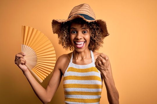 African American Tourist Woman With Curly On Vacation Wearing Summer Hat Using Hand Fan Screaming Proud And Celebrating Victory And Success Very Excited, Cheering Emotion