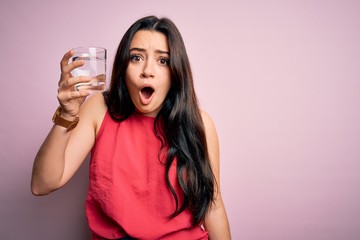 Young brunette woman drinking a glass of fresh water over pink isolated background scared in shock with a surprise face, afraid and excited with fear expression
