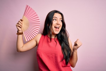 Young brunette woman holding hand fan for fresh air over pink isolated background pointing and showing with thumb up to the side with happy face smiling