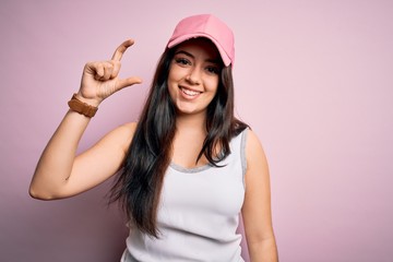 Obraz na płótnie Canvas Young brunette woman wearing casual sport cap over pink background smiling and confident gesturing with hand doing small size sign with fingers looking and the camera. Measure concept.