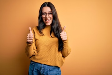 Young brunette woman wearing glasses and casual sweater over yellow isolated background success...
