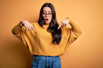 Young brunette woman wearing glasses and casual sweater over yellow isolated background Pointing down with fingers showing advertisement, surprised face and open mouth