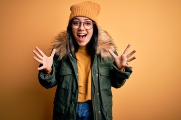 Young brunette woman wearing glasses and winter coat with hat over yellow isolated background celebrating crazy and amazed for success with arms raised and open eyes screaming excited. Winner concept