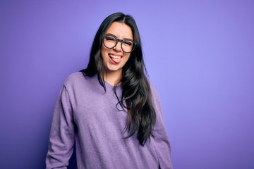 Young brunette woman wearing glasses over purple isolated background sticking tongue out happy with funny expression. Emotion concept.