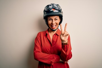 Middle age motorcyclist woman wearing motorcycle helmet over isolated white background smiling with happy face winking at the camera doing victory sign. Number two.