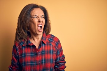 Middle age beautiful woman wearing casual shirt standing over isolated yellow background angry and mad screaming frustrated and furious, shouting with anger. Rage and aggressive concept.