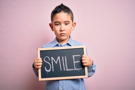 Young little boy kid showing blackboard with smile word as happy message over pink background with a confident expression on smart face thinking serious