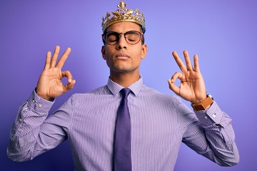 Young handsome african american man wearing golden crown of king over purple background relax and smiling with eyes closed doing meditation gesture with fingers. Yoga concept.