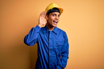 Young handsome african american worker man wearing blue uniform and security helmet smiling with hand over ear listening an hearing to rumor or gossip. Deafness concept.