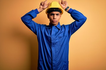 Young handsome african american worker man wearing blue uniform and security helmet doing funny gesture with finger over head as bull horns