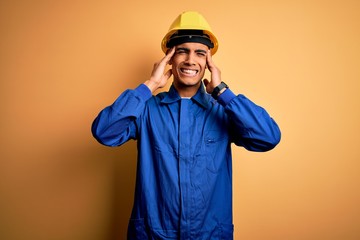 Young handsome african american worker man wearing blue uniform and security helmet covering ears with fingers with annoyed expression for the noise of loud music. Deaf concept.
