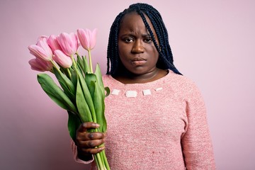 Young african american plus size woman with braids holding bouquet of pink tulips flower skeptic...