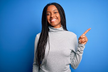 Young african american woman standing wearing casual turtleneck over blue isolated background with a big smile on face, pointing with hand finger to the side looking at the camera.